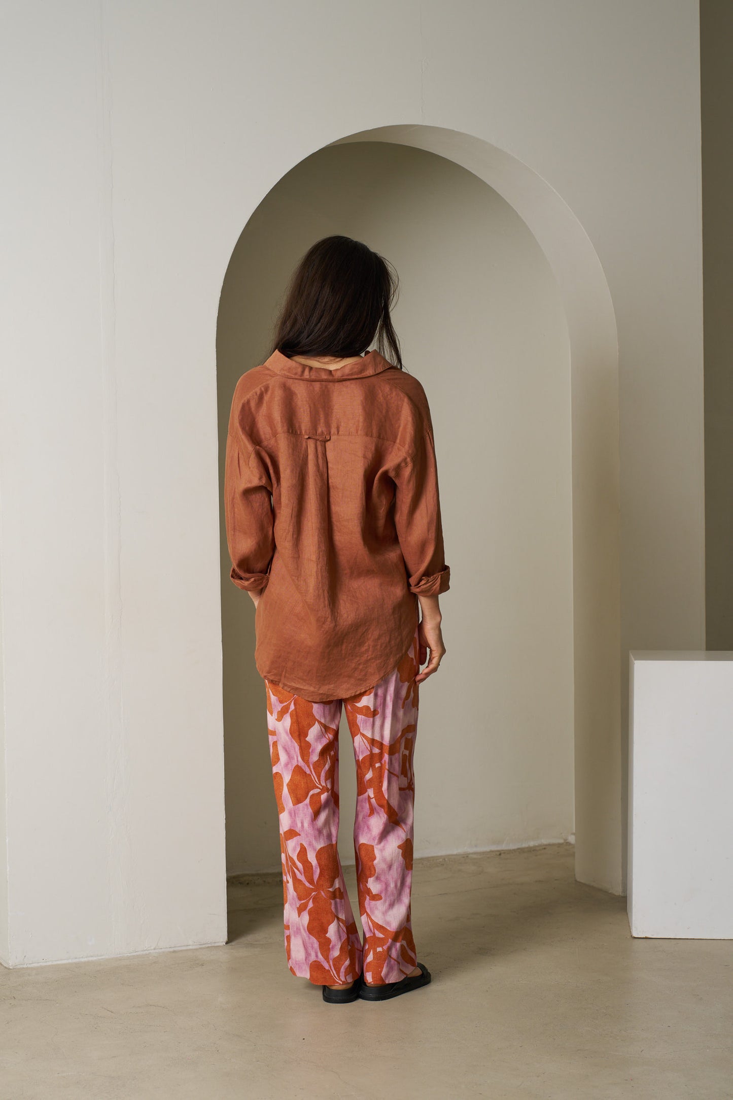 Person wearing a brown jacket and patterned pants standing in an archway.